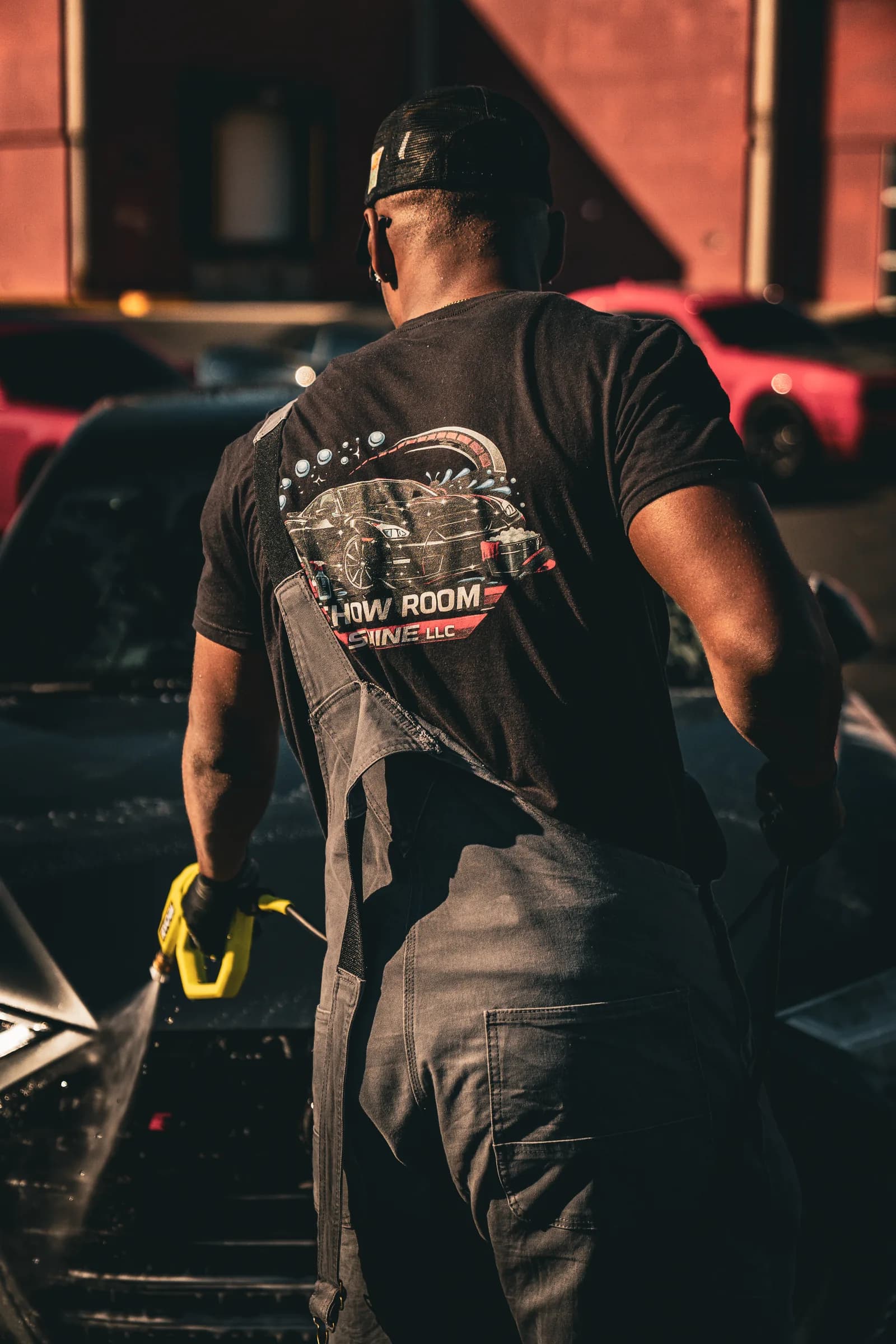 A Showroom Shine detailer pressure-washing a car at golden hour, the company logo across his back